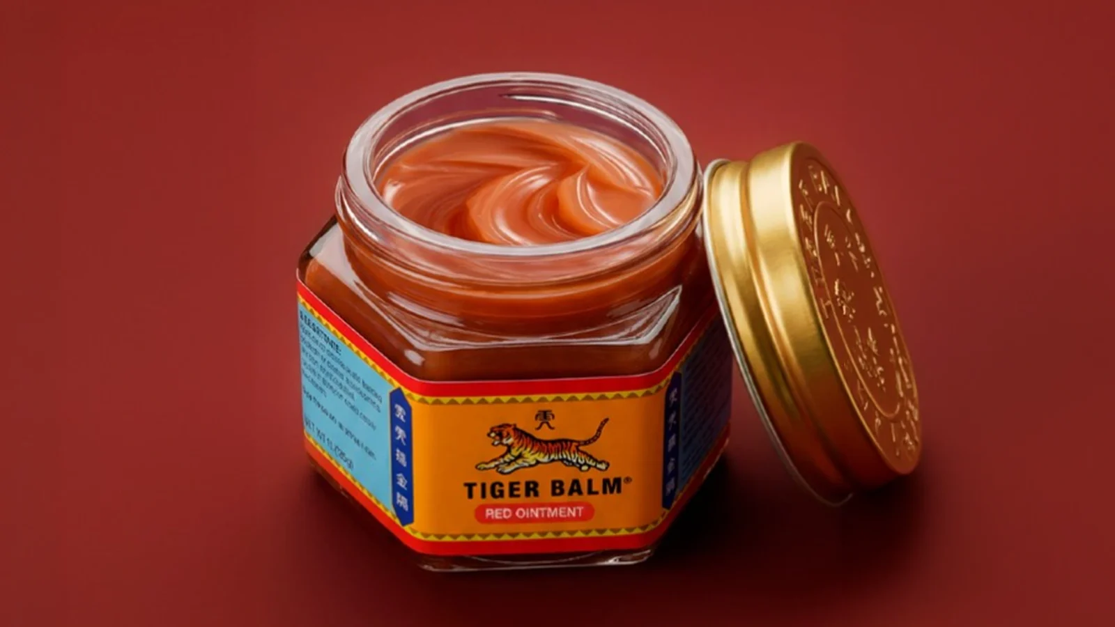 Pot hexagonal de Tiger Balm Vermell obert sobre fons vermell, mostrant l'ungüent i la tapa daurada